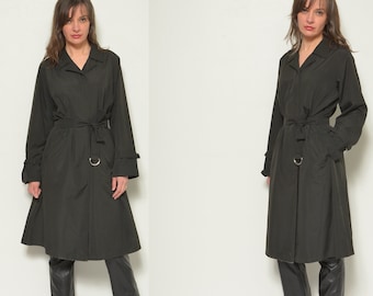 May include: A dark green trench coat with a collared neckline, long sleeves, and a belted waist. The coat features a knee-length design and is paired with black pants and boots.
