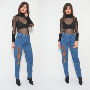 May include: Blue denim jeans with brown decorative stitching on the front. The jeans have a high waist and a straight leg. The model is wearing a black sheer long-sleeve top and black ankle boots.