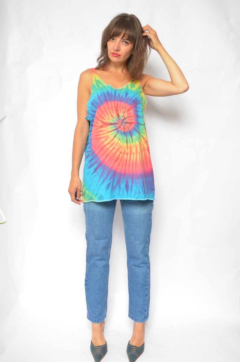 Puede incluir: Una mujer con un top de tirantes tie-dye con un patr&oacute;n de espiral azul, rosa, amarillo y verde. Tambi&eacute;n lleva pantalones vaqueros azules y zapatos azul oscuro.