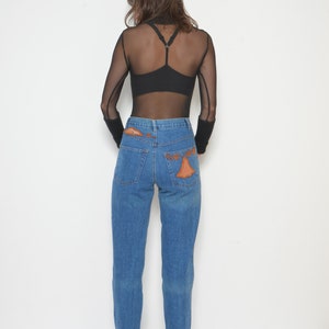 May include: A person wearing a black mesh long-sleeve top and blue jeans. The jeans have orange decorative patches on the back pockets. The person is also wearing black heeled shoes.