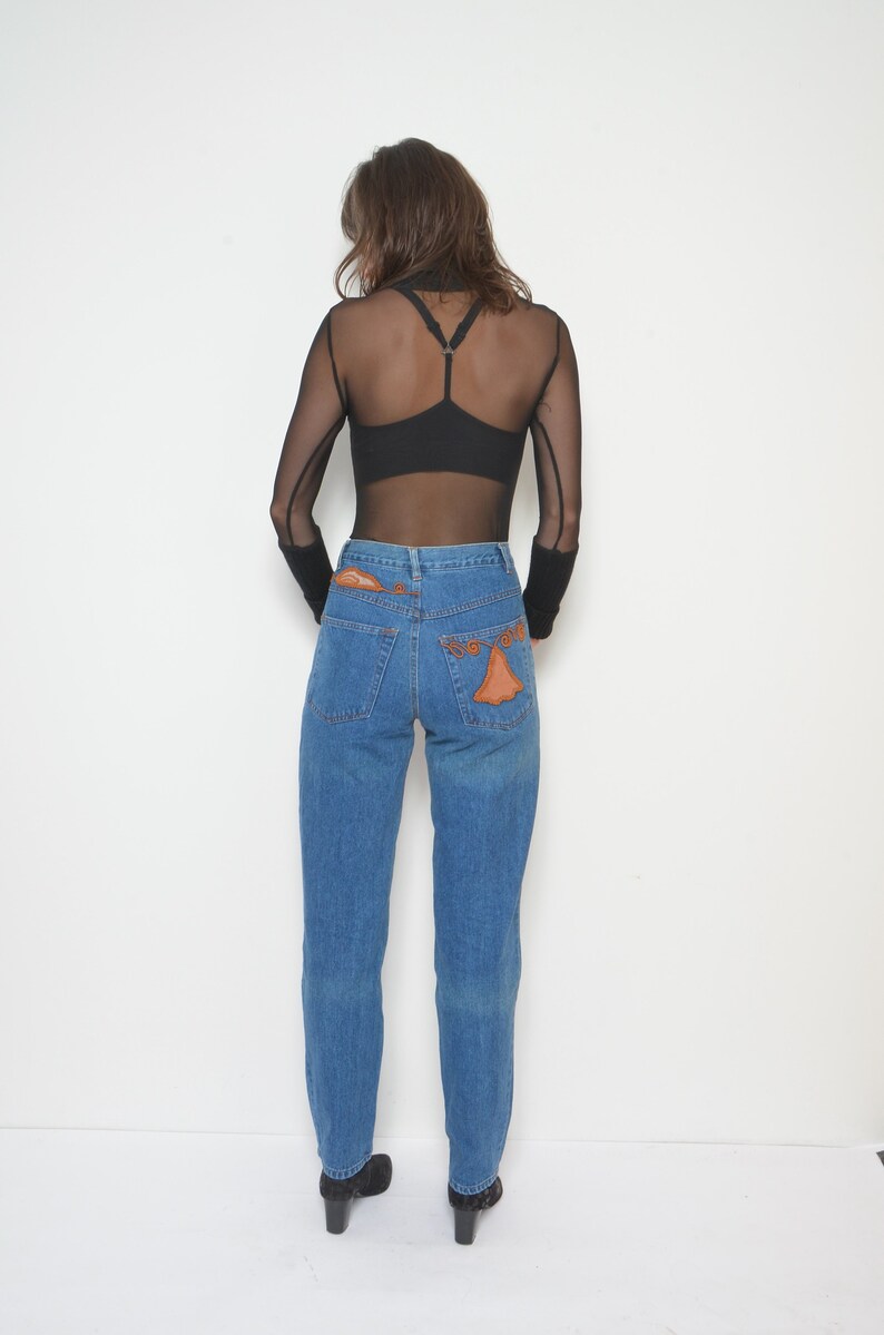 May include: A person wearing a black mesh long-sleeve top and blue jeans. The jeans have orange decorative patches on the back pockets. The person is also wearing black heeled shoes.