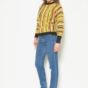May include: A woman wearing a green, orange, and black striped sweater with a black collar and long sleeves. She is also wearing blue jeans and black ankle boots.