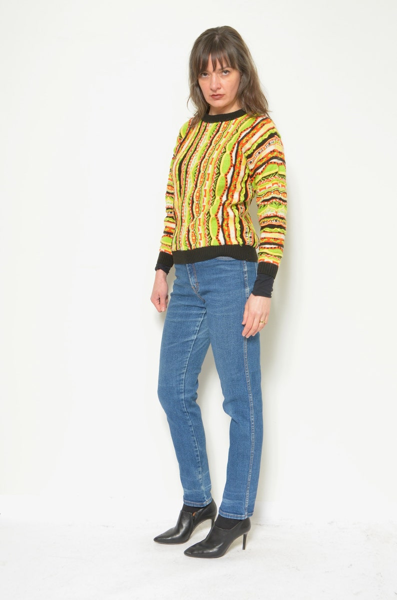 May include: A woman wearing a green, orange, and black striped sweater with a black collar and long sleeves. She is also wearing blue jeans and black ankle boots.