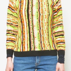 May include: A colorful striped sweater with a black crew neck. The sweater has a geometric pattern in shades of green, orange, black, and white.