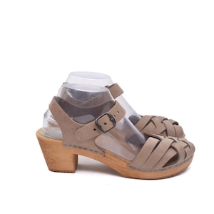 May include: A pair of beige sandals with a wooden heel. The sandals have a woven design across the toes and a buckle closure on the ankle strap. The heel is approximately 7.6 cm high.