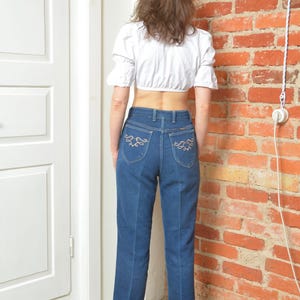 May include: A white crop top with short sleeves and blue jeans. The jeans have decorative stitching on the back pockets. The person is wearing brown and gold boots.