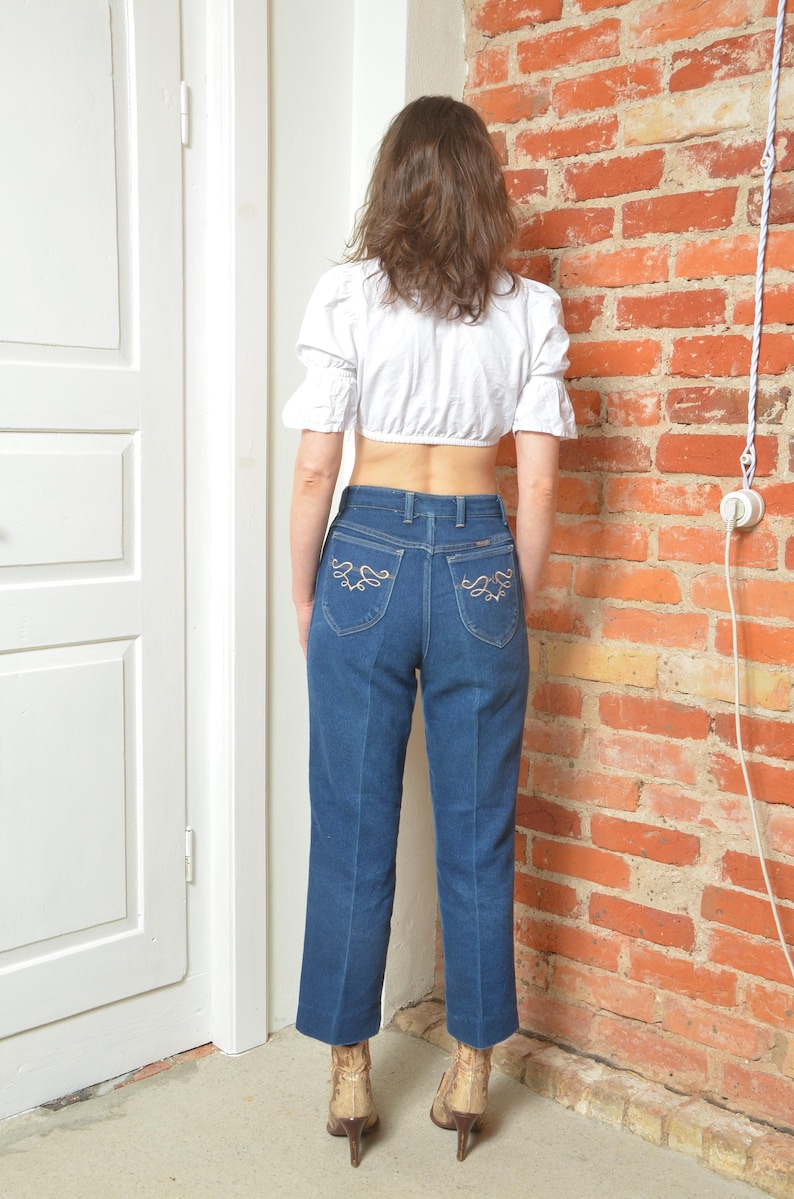May include: A white crop top with short sleeves and blue jeans. The jeans have decorative stitching on the back pockets. The person is wearing brown and gold boots.