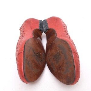 May include: Pair of red leather shoes with a woven design on the upper. The soles are brown with wear and the heels are black. The shoes are angled towards the viewer, showcasing the soles and heels.