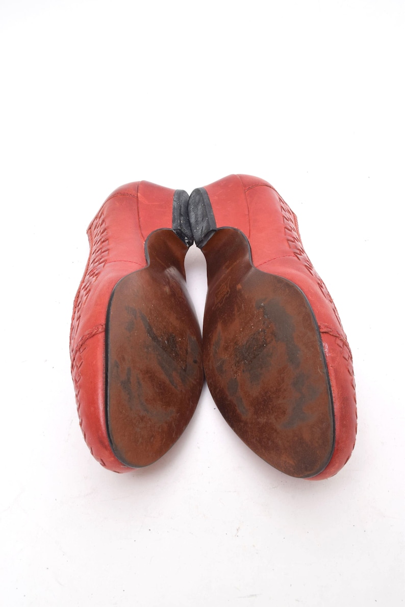 May include: Pair of red leather shoes with a woven design on the upper. The soles are brown with wear and the heels are black. The shoes are angled towards the viewer, showcasing the soles and heels.