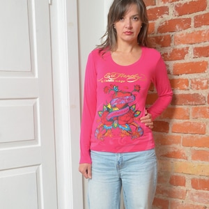 May include: A long-sleeved, hot pink shirt with a scoop neck. The shirt features a colorful graphic design with roses, a heart, and the text "Ed Hardy" in gold lettering. The model is wearing light blue jeans.