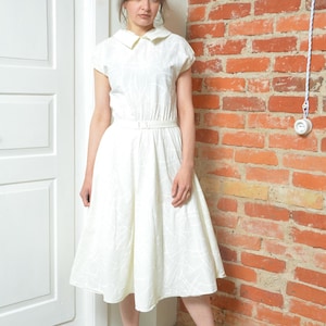 Può includere: Un abito bianco in stile vintage con collo e maniche corte. L'abito presenta un corpetto aderente, una vita con cintura e una gonna svasata. L'abito è abbinato a tacchi arancioni. Lo sfondo comprende un muro di mattoni e una porta bianca.