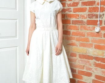 May include: A white, vintage-style dress with a collared neckline and cap sleeves. The dress features a fitted bodice, a belted waist, and a flared skirt. The dress is paired with orange heels. The background includes a brick wall and a white door.