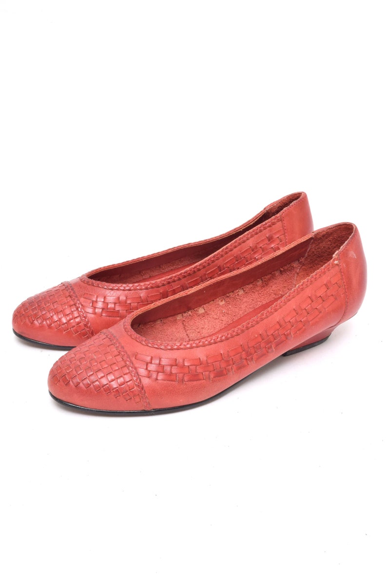 May include: A pair of coral-colored leather ballet flats. The toe box features a woven design, and the shoes have a low heel. The shoes are set against a white background.