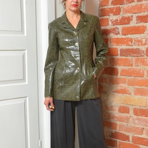 Może przedstawiać: Zielony, oliwkowy żakiet ze sztucznej skóry z wzorem skóry węża. Żakiet ma kołnierz z klapami, dwie przednie kieszenie i trzy guziki. Modelka ma na sobie czarne spodnie z szerokimi nogawkami. Tło to ceglana ściana i białe drzwi.