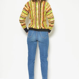 May include: A woman wearing a green, orange, and brown striped sweater with a black turtleneck and blue jeans.