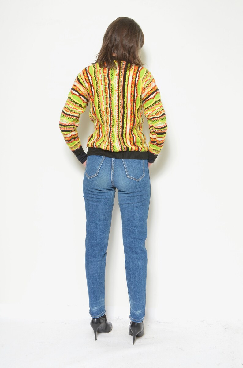 May include: A woman wearing a green, orange, and brown striped sweater with a black turtleneck and blue jeans.