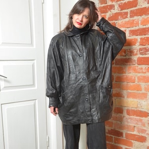 May include: A black leather jacket with a button-up front, two pockets, and a high collar. The jacket has a slightly oversized fit and is worn over a black turtleneck. The model is standing in front of a brick wall and a white door.