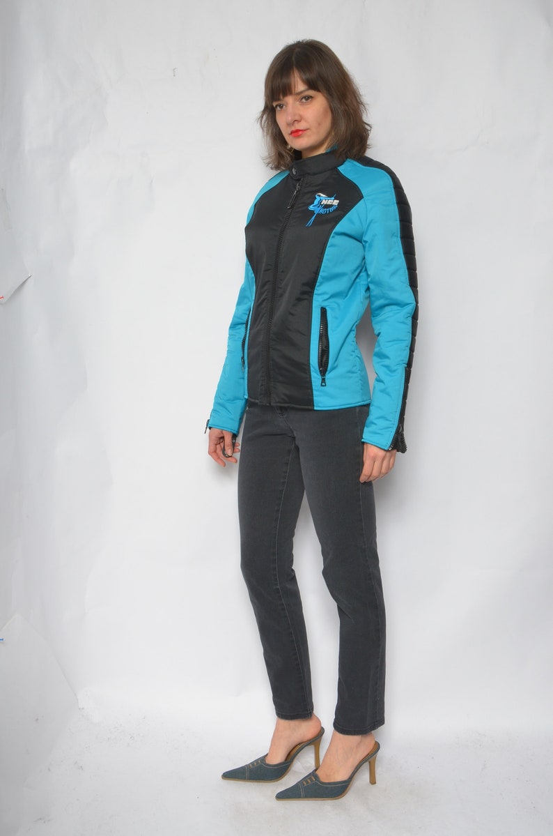May include: A black and blue jacket with a zipper closure and a logo on the left side. The jacket has a quilted design on the sleeves. The person is wearing black jeans and blue heeled shoes.