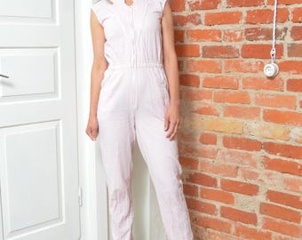 May include: A sleeveless, light pink jumpsuit with a subtle checkered pattern. The jumpsuit features a zip-up front, a stand-up collar, and fitted legs. The model is wearing orange heels. The background includes a white door and a red brick wall.