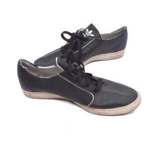 May include: A pair of black leather Adidas trainers with white laces and a white Adidas logo on the side. The shoes have a white rubber sole.