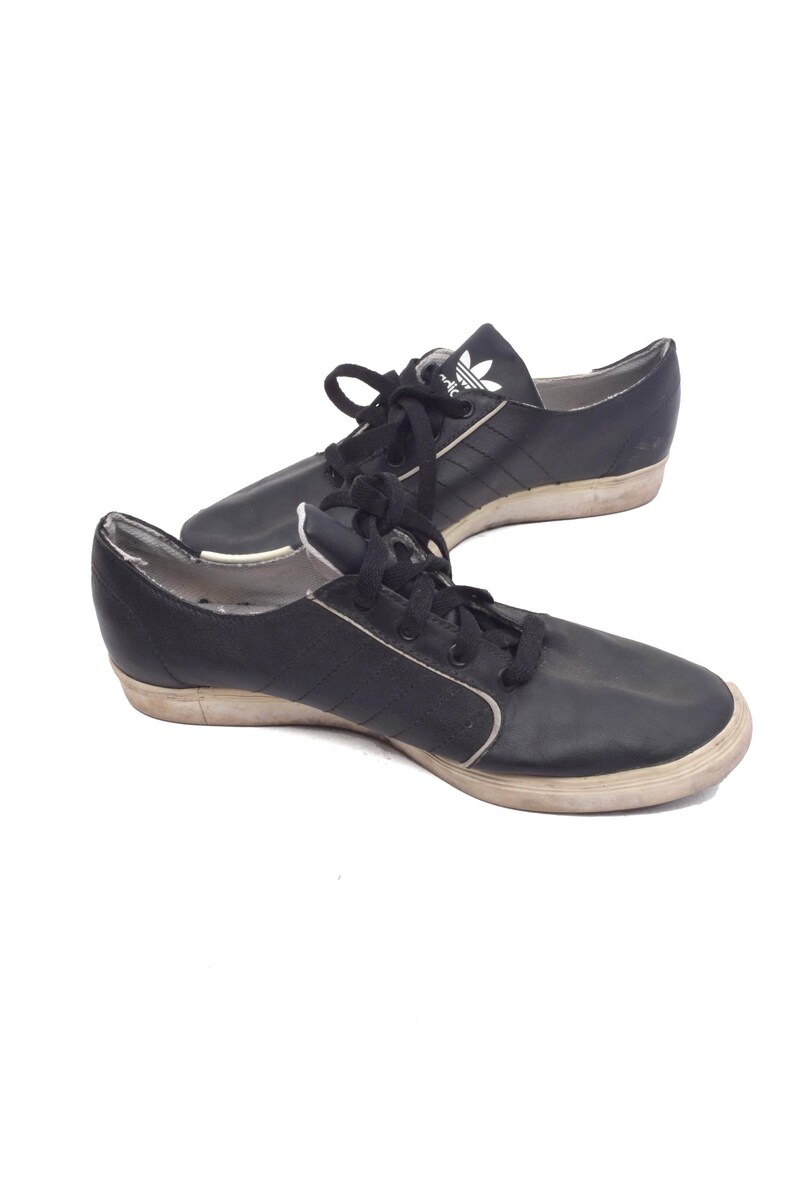 May include: A pair of black leather Adidas trainers with white laces and a white Adidas logo on the side. The shoes have a white rubber sole.