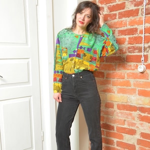Pode incluir: Blusa de manga comprida com um padrão colorido de formas geométricas verdes, amarelas, roxas e laranja. A blusa está abotoada. A pessoa está usando jeans pretos.