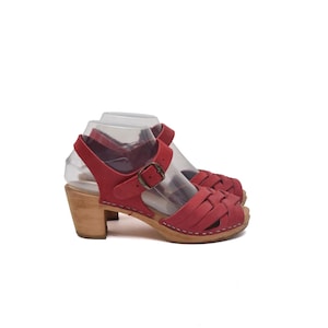May include: A pair of red leather clogs featuring a woven strap design across the front, an adjustable ankle strap with a metal buckle, and a light-coloured wooden block heel. The shoes are displayed on clear plastic forms against a white background.