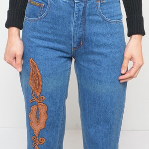 May include: Blue denim jeans with orange stitching and a decorative brown leather and embroidered design on the left leg. The jeans have a classic five-pocket design and a button closure.
