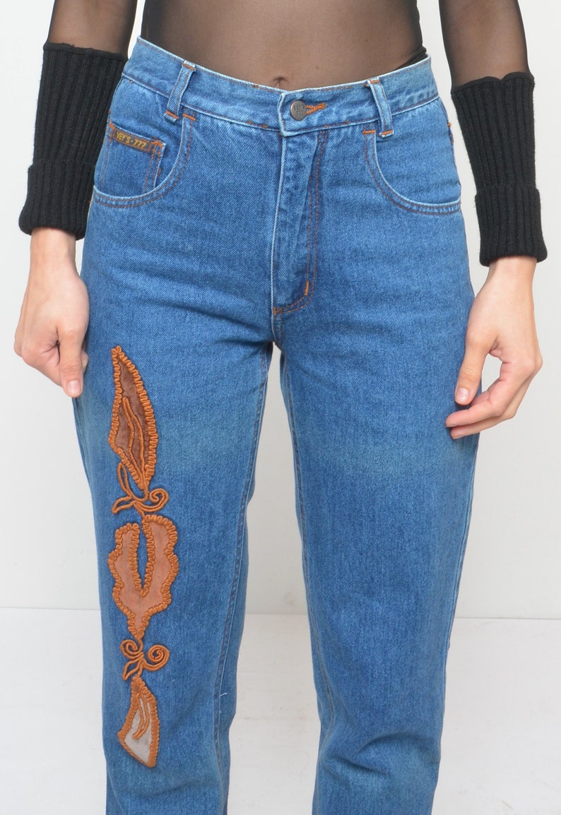 May include: Blue denim jeans with orange stitching and a decorative brown leather and embroidered design on the left leg. The jeans have a classic five-pocket design and a button closure.