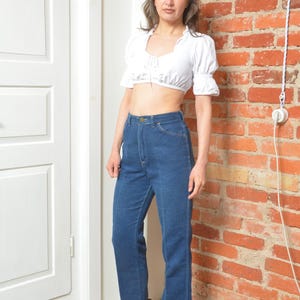 May include: A white crop top with short, puffed sleeves and a tie-up front is worn with high-waisted, dark blue denim jeans. The outfit is completed with patterned ankle boots. The background features a brick wall and a white door.