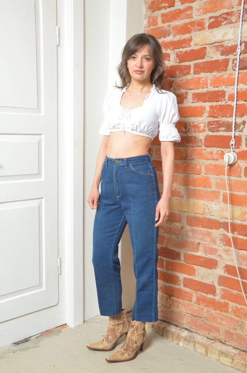 May include: A white crop top with short, puffed sleeves and a tie-up front is worn with high-waisted, dark blue denim jeans. The outfit is completed with patterned ankle boots. The background features a brick wall and a white door.