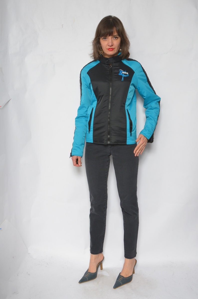 May include: A black and blue jacket with a zipper closure and a logo on the chest. The jacket is worn by a person with dark hair and is paired with black jeans and blue shoes.
