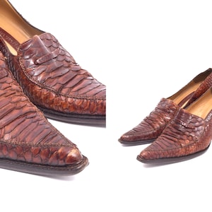 Pode incluir: Um par de sapatos de salto alto de pele de cobra castanho com bico fino. Os sapatos têm um design de deslizamento e um salto baixo.