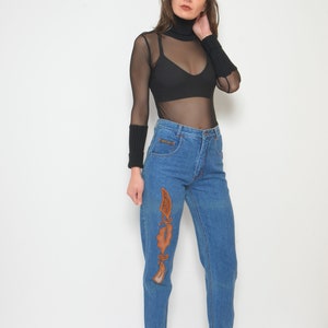 May include: A woman wearing a black mesh long-sleeve top and high-waisted blue jeans. The jeans feature an embroidered design in shades of brown. She is also wearing black ankle boots.