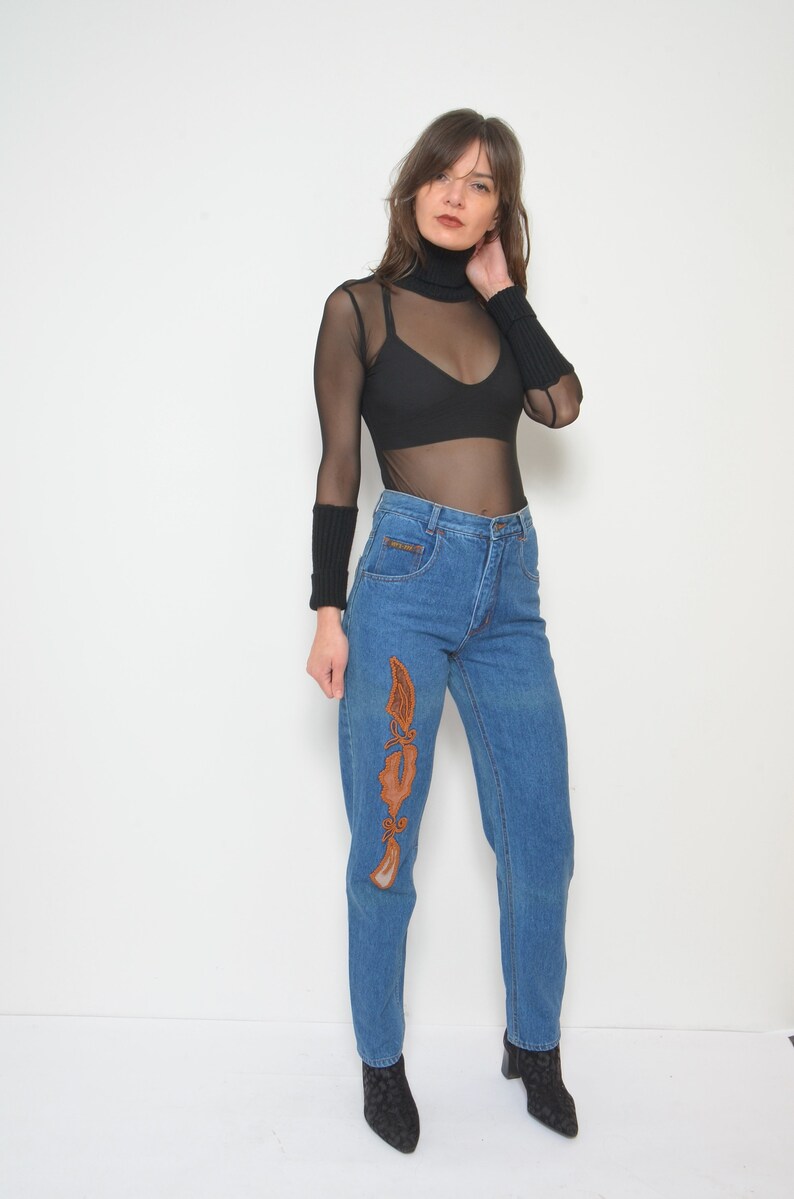 May include: A woman wearing a black mesh long-sleeve top and high-waisted blue jeans. The jeans feature an embroidered design in shades of brown. She is also wearing black ankle boots.