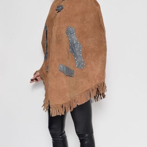 May include: A brown suede poncho with fringe and leather accents. The poncho has a high collar and is worn over black leggings and black wedge boots.