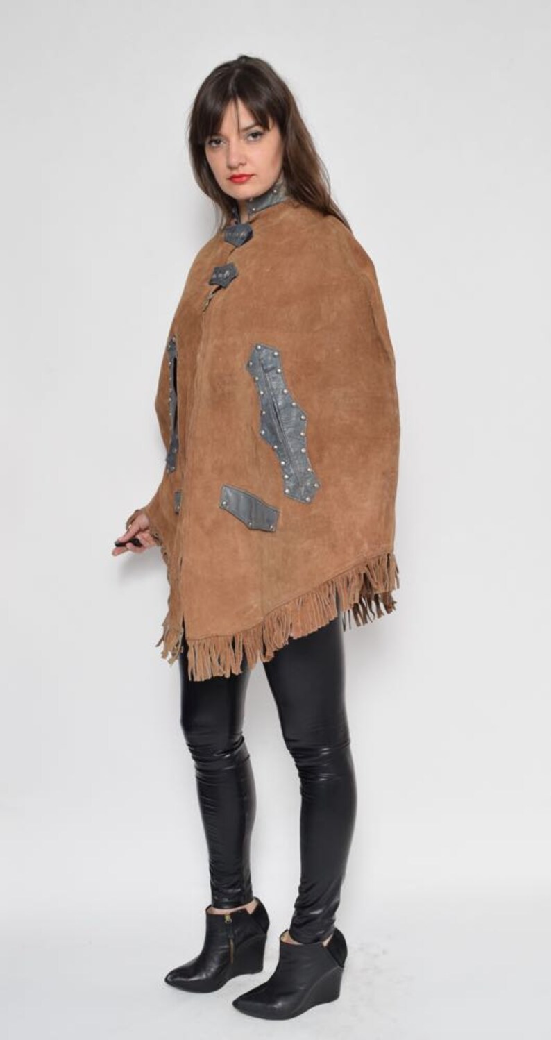 May include: A brown suede poncho with fringe and leather accents. The poncho has a high collar and is worn over black leggings and black wedge boots.