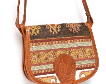 May include: A brown leather crossbody bag with a colorful woven fabric flap. The flap features a geometric pattern in shades of brown, red, yellow, and white. A leather logo is embossed on the front of the flap.