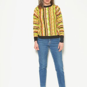 May include: A woman wearing a green, orange, and black striped sweater with a black trim and blue jeans. She is wearing black heeled boots.