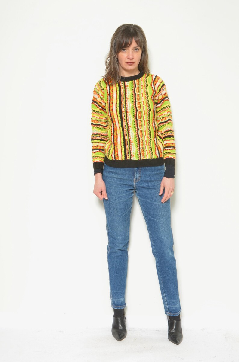 May include: A woman wearing a green, orange, and black striped sweater with a black trim and blue jeans. She is wearing black heeled boots.