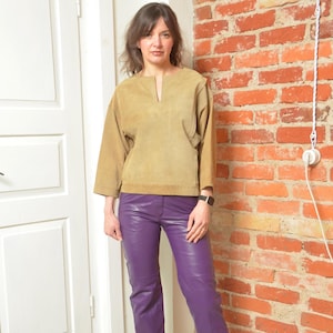 May include: A tan suede top with a V-neck and three-quarter sleeves paired with purple leather pants. The outfit is worn by a person standing in front of a brick wall and a white door.