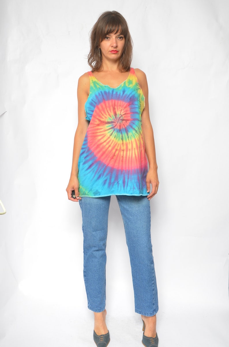 Puede incluir: Una mujer con un top de tirantes tie-dye con un patr&oacute;n de espiral arco&iacute;ris y pantalones vaqueros azules.