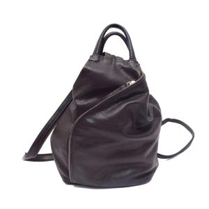 Peut inclure: Un sac à dos en cuir marron foncé avec une silhouette souple et affaissée, doté d'une poignée supérieure, d'une poche frontale zippée en diagonale et de fines bretelles réglables. Le sac présente une base arrondie et une finition texturée.