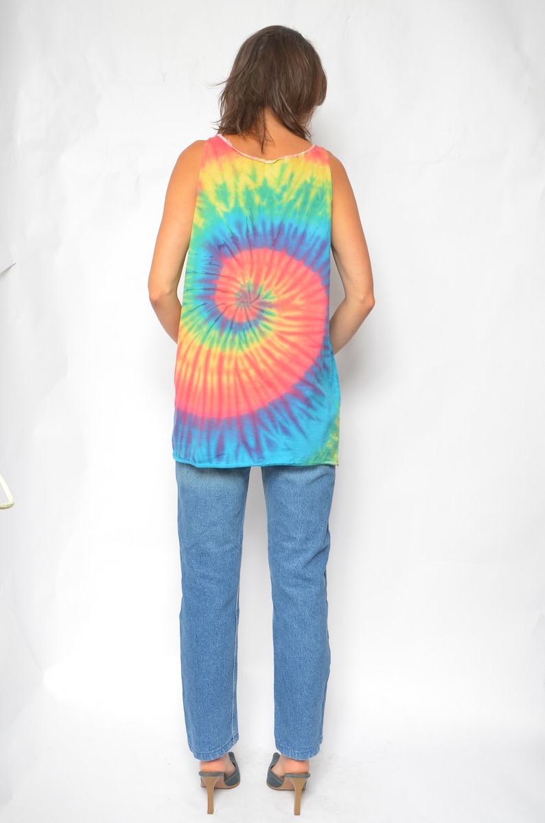 Puede incluir: Una mujer con un top sin mangas de tie-dye con un patr&oacute;n de espiral azul, rosa, amarillo y verde. Tambi&eacute;n lleva pantalones vaqueros azules y zapatos de tac&oacute;n negros.