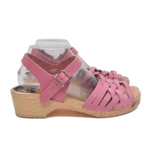 Puede incluir: Un par de sandalias de cuero rosa con un diseño de tiras trenzadas en la parte delantera, con una correa de tobillo ajustable con hebilla de metal plateado y una suela de madera clara con tacón bajo, presentadas sobre un pie de plástico.