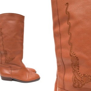 Puede incluir: Un par de botas altas de cuero marrón. Las botas tienen un diseño bordado decorativo en el lateral. Tienen un tacón bajo y una puntera redondeada. Las botas están hechas de cuero liso y texturizado.
