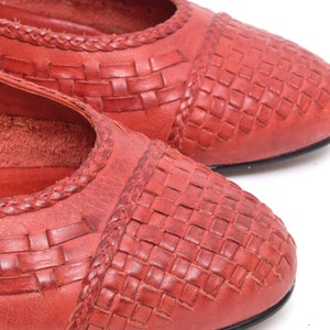 May include: Close-up of two red leather flats. The shoes have a woven pattern on the toe and a braided trim along the top edge. The soles are black. The shoes are on a white background.