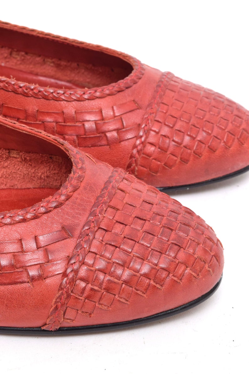 May include: Close-up of two red leather flats. The shoes have a woven pattern on the toe and a braided trim along the top edge. The soles are black. The shoes are on a white background.