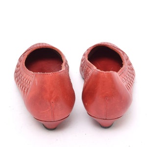 May include: A pair of red leather ballet flats. The shoes have a woven texture on the top and a small heel. The shoes are displayed on a white background.