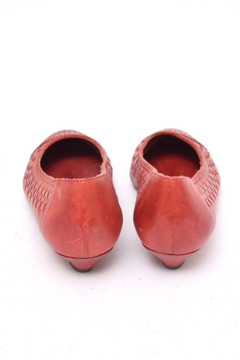 May include: A pair of red leather ballet flats. The shoes have a woven texture on the top and a small heel. The shoes are displayed on a white background.